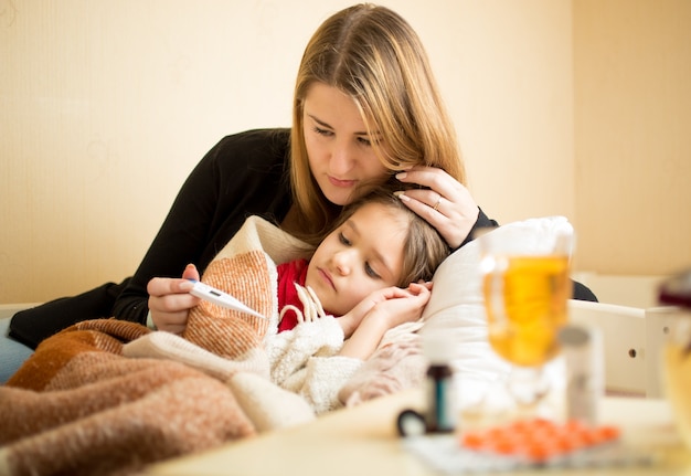 Joven madre comprobando la temperatura de la hija enferma acostada en la cama