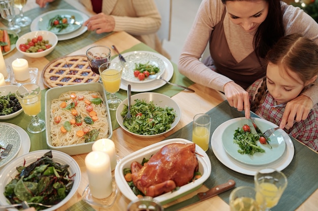 Joven madre cortando tomate en el plato de su pequeña hija mientras ambos están sentados junto a la mesa festiva y cenando