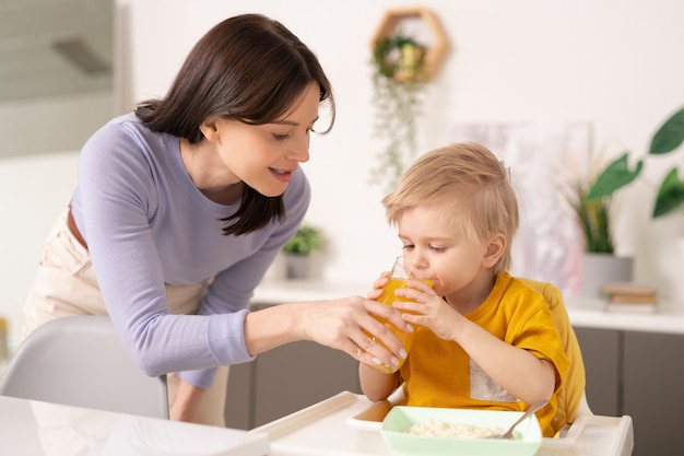 Joven madre cuidadosa sosteniendo un vaso de jugo de naranja mientras se inclina por su pequeño y lindo hijo bebiéndolo en una pequeña mesa durante el desayuno