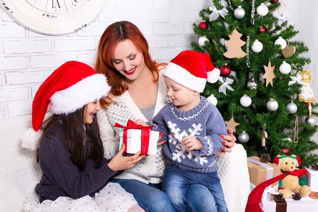 Joven madre dando regalos a sus hijos delante del árbol de Navidad