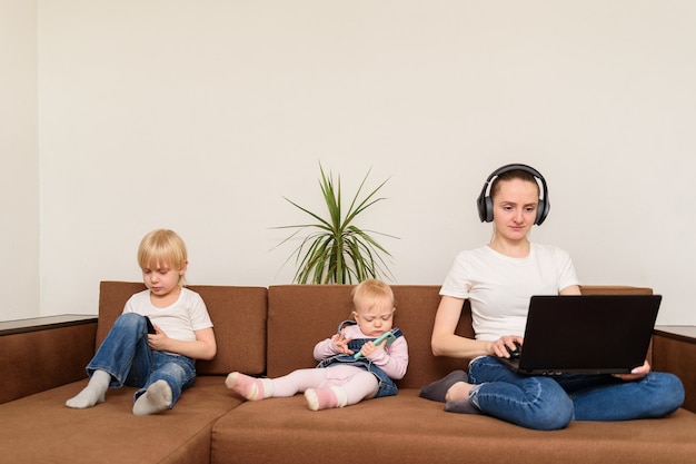 Joven madre y dos niños sentados en el sofá con la computadora y el teléfono. Adicción a las personas y la tecnología