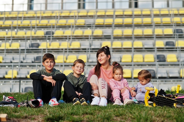 Joven madre elegante con cuatro hijos sentados en el césped contra el estadio La familia deportiva pasa tiempo libre al aire libre con scooters y patines