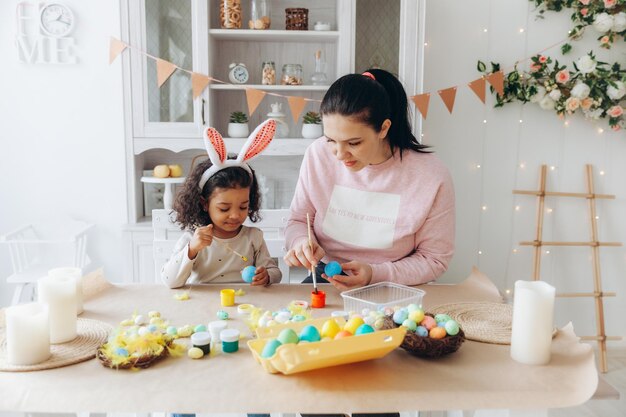 Joven madre europea enseñando a un niño afroamericano cómo pintar y decorar huevos con pinturas para las vacaciones de pascua sentados juntos en la mesa de la cocina