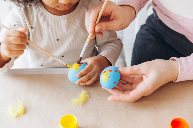 Joven madre europea enseñando a un niño afroamericano cómo pintar y decorar huevos con pinturas para las vacaciones de pascua sentados juntos en la mesa de la cocina