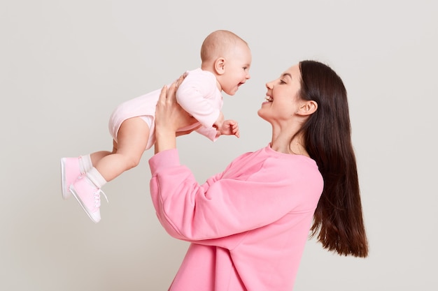 Foto joven madre europea sosteniendo a su bebé en las manos y mirando a su bebé con amor y sonrisa suave, mujer con cabello oscuro con suéter casual rosa, jugando con su hijo.