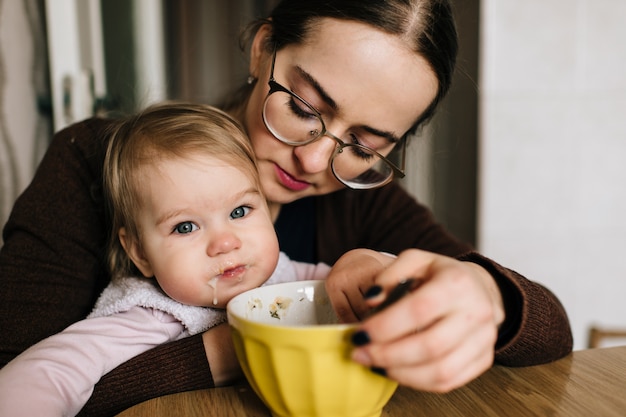 Joven madre feliz alimentando a su pequeño bebé en la mesa.