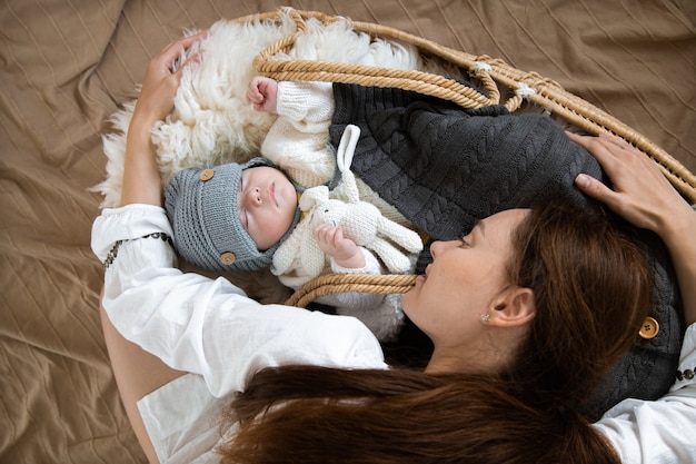 Joven madre feliz y un bebé durmiendo en una cuna de mimbre en un gorro de punto caliente debajo de una manta caliente con un juguete en el mango.