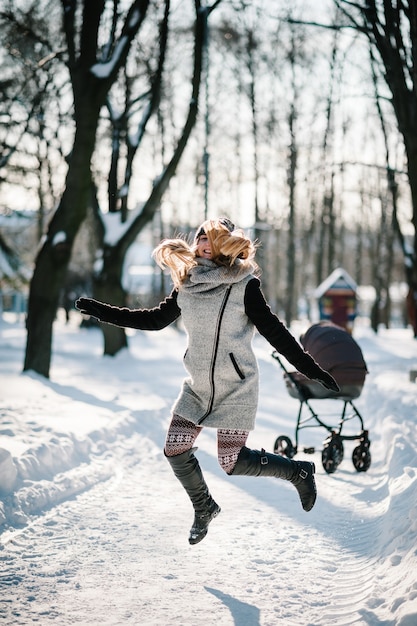 Una joven madre feliz salta, camina con un cochecito y un bebé en un parque de invierno