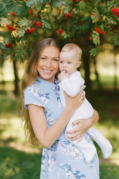 Una joven madre feliz sostiene a su pequeño hijo en sus brazos contra el fondo de un árbol Rowan con bayas rojas en el verano en un parque o jardín y sonríe. Día de la Madre