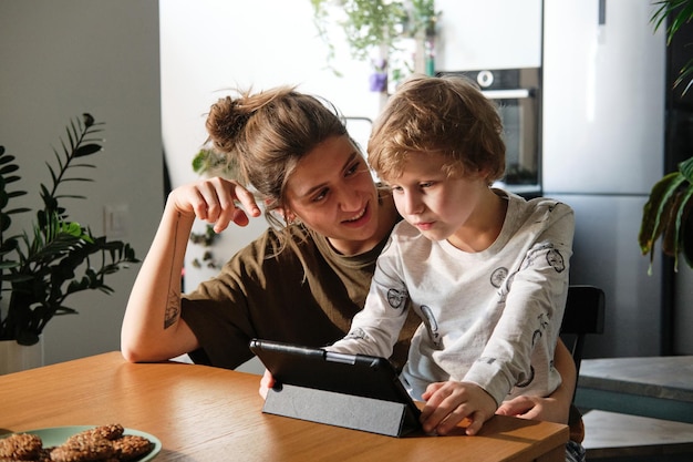 Joven madre hablando con su hijo mientras juega tableta digital en la mesa de la cocina