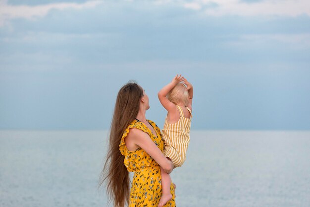 Joven madre hermosa sostiene a su pequeña hija y mira el mar Amor de madre