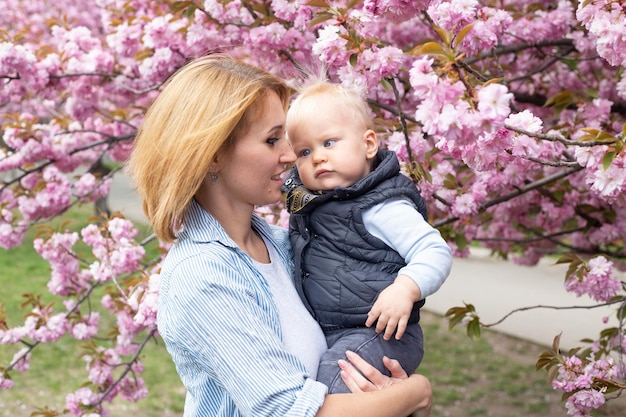 Joven madre con un hijo pequeño en sus manos en el parque con el árbol de flor de cerezo de Sakura Feliz madre e hijo