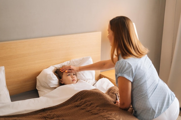 La joven madre mide la temperatura toca la frente de la hija enferma acostada en la cama en casa. Padres que cuidan a un niño enfermo acostado en la cama con gripe. Concepto de salud infantil.