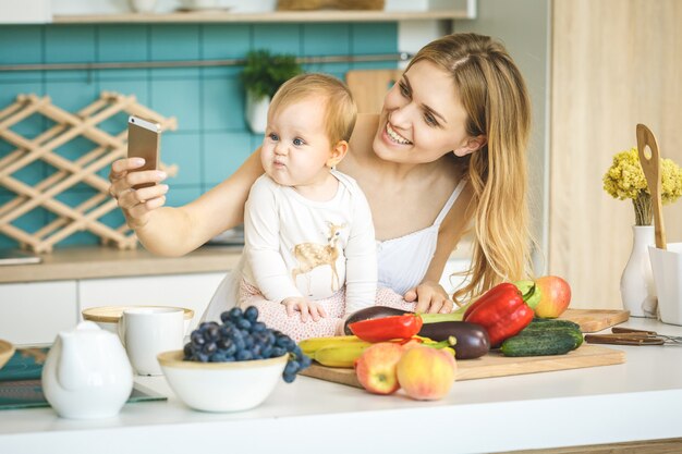 Joven madre mirando a cámara y sonriendo, cocinando y jugando con su pequeña hija en una cocina moderna.