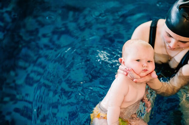 Joven madre, niña feliz en la piscina. Enseña a los bebés a nadar. Disfruta el primer día de natación en el agua. Mamá sostiene al niño preparándose para bucear. haciendo ejercicios. mano que lleva al niño en el agua