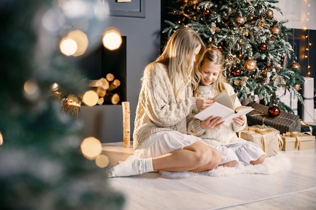 Joven madre y niña sentada árbol de navidad en casa y leyendo un libro