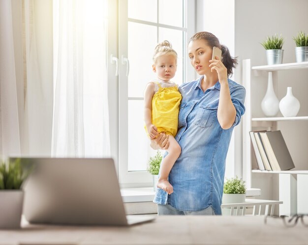 Joven madre con niño pequeño trabajando en la computadora, hablando por teléfono en casa.