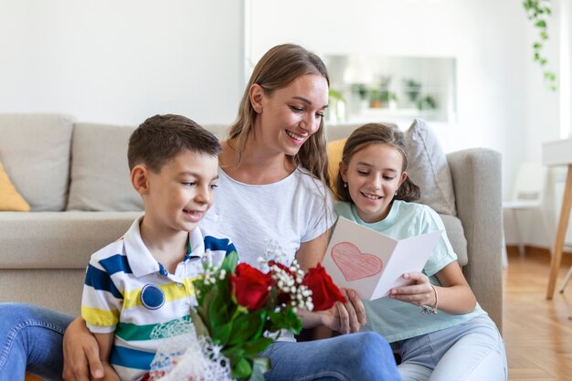 Joven madre con un ramo de rosas se ríe, abrazando a su hijo, y una niña alegre con una tarjeta y rosas felicita a mamá durante la celebración navideña en la cocina de casa. día de la Madre