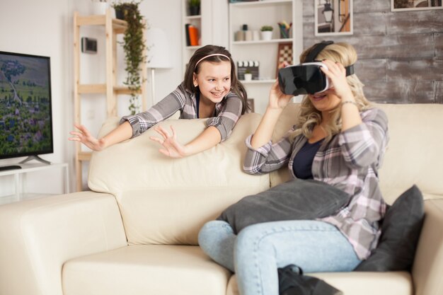 Joven madre sentada en el sofá de la sala de estar con gafas de realidad virtual. Hija alegre con tirantes.