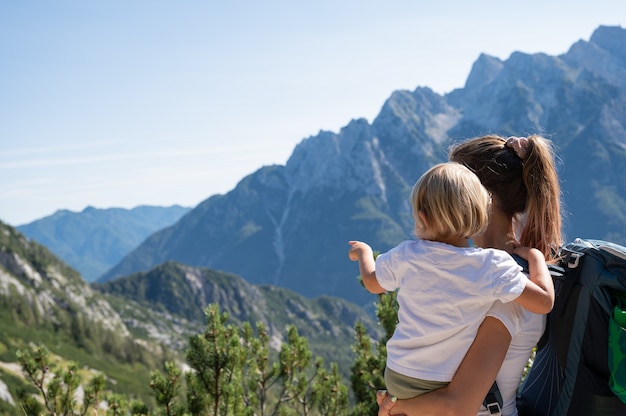 Joven madre sosteniendo a su hija pequeña mirando la hermosa vista de las montañas mientras caminan juntos en un día soleado.