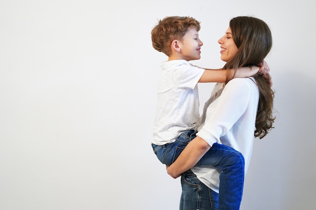 Joven madre sosteniendo a su hijo preadolescente sonriendo y mirándolo a los ojos en la pared blanca con espacio de copia