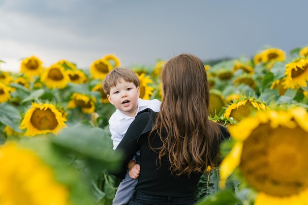 Una joven madre sostiene a su hijo en brazos caminando por un campo con girasoles en un día de verano
