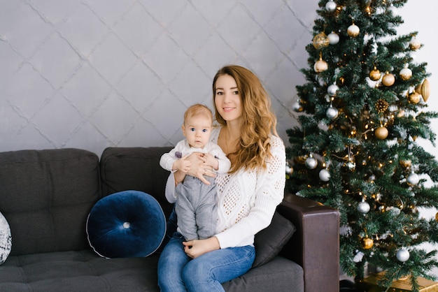 Joven madre sostiene a su pequeño hijo en sus brazos cerca del árbol de Navidad en la sala de estar