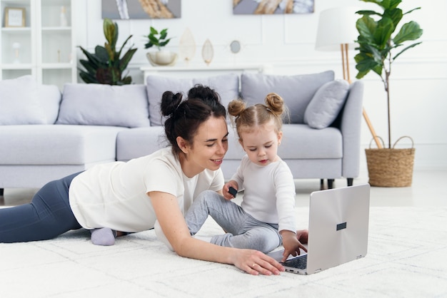 Joven madre y su adorable hija están usando la computadora portátil mientras está acostado en el piso.