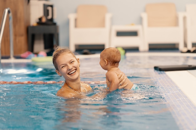 Joven madre y su bebé disfrutando de una lección de natación para bebés en la piscina. Niño divirtiéndose en el agua con mamá