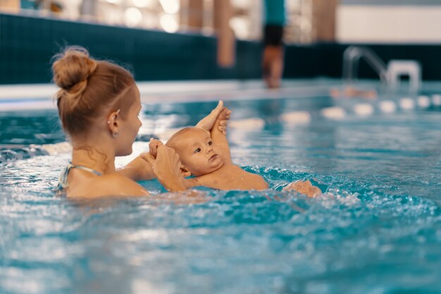 Joven madre y su bebé disfrutando de una lección de natación para bebés en la piscina. Niño divirtiéndose en el agua con mamá