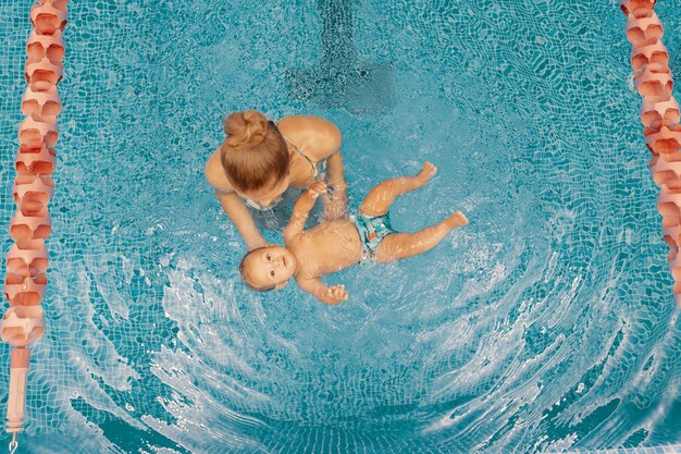 Joven madre y su bebé disfrutando de una lección de natación para bebés en la piscina. Niño divirtiéndose en el agua con mamá