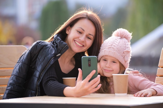 La joven madre y su hija se toman una foto con la cámara selfie del teléfono sentados en un café de la calle con bebidas calientes en el soleado día de otoño Presencia en las redes sociales en el concepto de la vida cotidiana