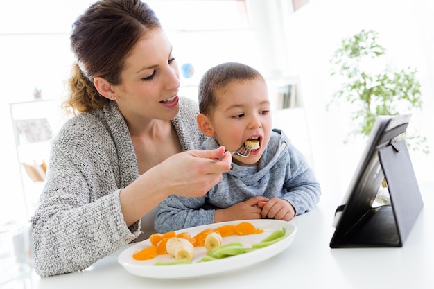 Joven madre y su hijo utilizando tableta digital mientras come frutas en casa.