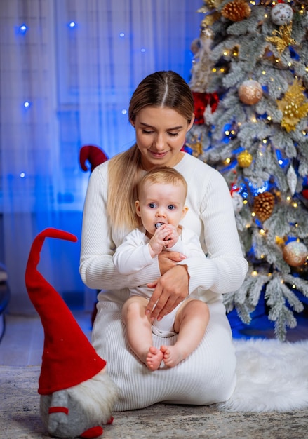 Una joven madre y su pequeña hija están sentadas en el suelo cerca del árbol de Navidad y sonriendo