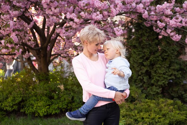 Joven madre y su pequeño hijo divirtiéndose en el parque de primavera cerca del árbol floreciente de sakura rosa Concepto de primavera