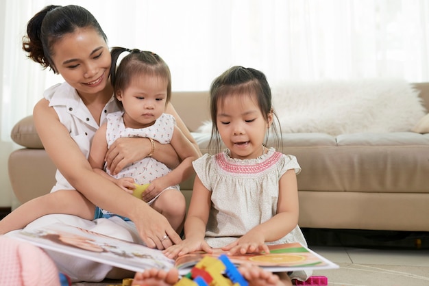 Joven madre y sus dos pequeñas hijas mirando coloridas imágenes en el libro