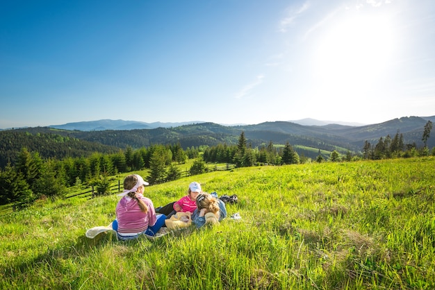 Foto la joven madre y sus hijas pequeñas se acuestan en una pendiente cubierta de hierba o admiran la hermosa vista de un zorro que crece en las colinas en un cálido día de verano. conceptos de camping y trekking. copyspace