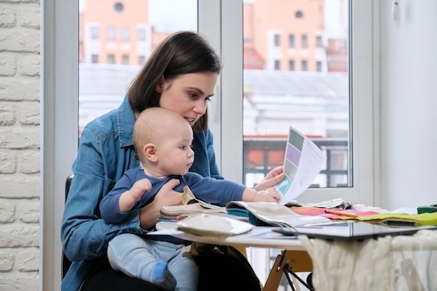 La joven madre trabaja con el bebé en brazos en la oficina en casa. Mujer diseñadora de interiores que trabaja con muestras de tela, en la tableta de mesa, bocetos y paletas textiles