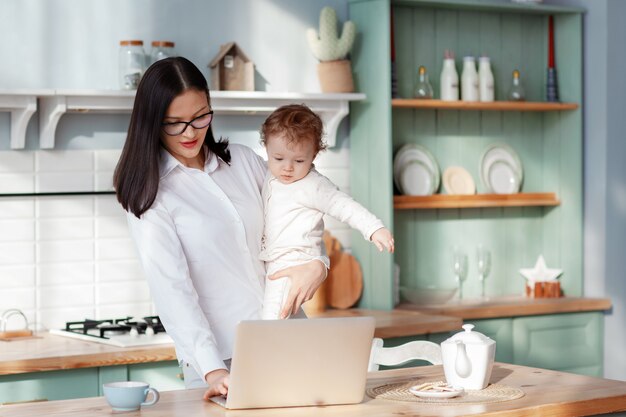 Foto joven madre trabaja en casa con una computadora portátil con un bebé en brazos