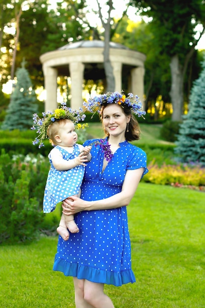 Joven madre con un vestido azul con lunares y una corona de flores silvestres con bebé en brazos.