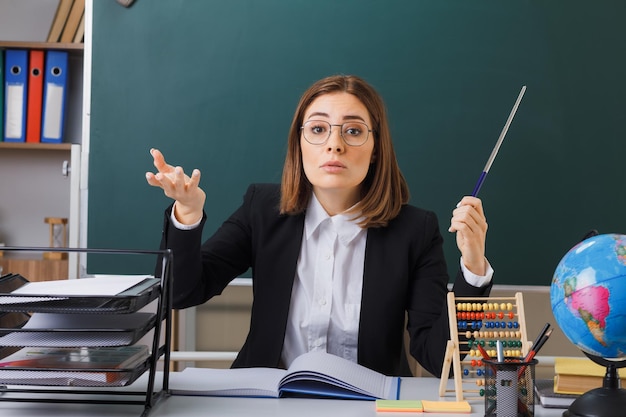 joven maestra con anteojos sentada en el escritorio de la escuela frente a la pizarra en el aula con ábaco y globo comprobando el registro de clase sosteniendo un puntero que parece disgustado levantando el brazo con disgusto