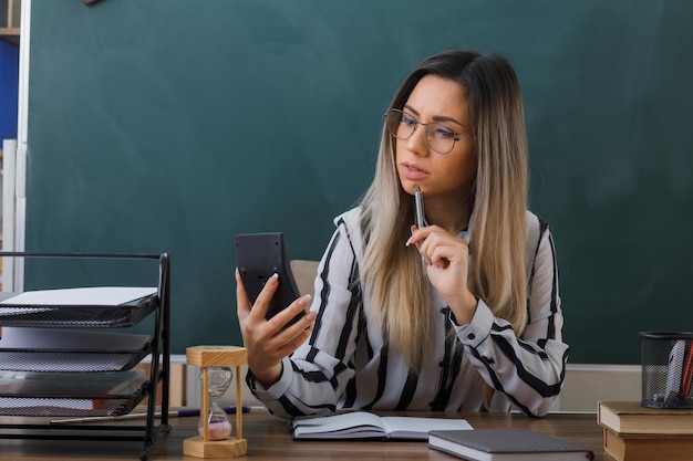 joven maestra con anteojos sentada en el escritorio de la escuela frente a la pizarra en el aula explicando la lección sosteniendo la calculadora mirándola desconcertada