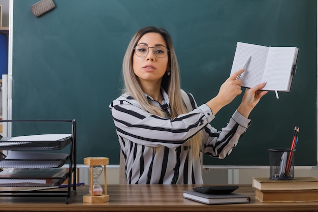 joven maestra con anteojos sentada en el escritorio de la escuela frente a la pizarra en el aula revisando la tarea de los estudiantes explicando la lección con confianza