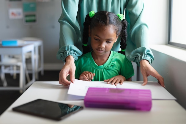 Joven maestra caucásica ayudando a una niña afroamericana de primaria que estudia en el escritorio