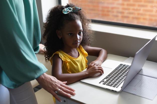 Una joven maestra caucásica de pie junto a una niña afroamericana que usa una computadora portátil en clase.
