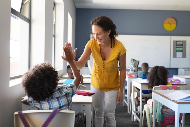 Una joven maestra caucásica sonriente dándole cinco a un niño afroamericano sentado en el escritorio.