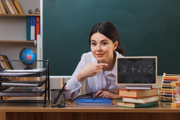 joven maestra complacida sosteniendo y apuntando a la mini pizarra sentada en el escritorio con herramientas escolares en el aula