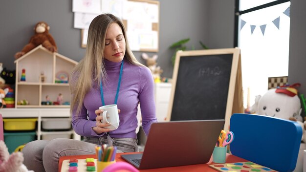 Joven maestra de preescolar rubia usando una laptop bebiendo café en el jardín de infantes