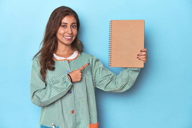 Joven maestra de primaria sosteniendo un cuaderno en un estudio azul