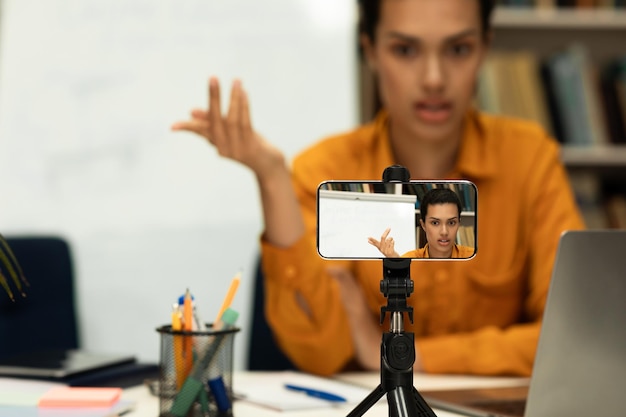 Joven maestra que brinda lecciones en línea mirando la cámara del teléfono celular o grabando video en un teléfono inteligente en un trípode